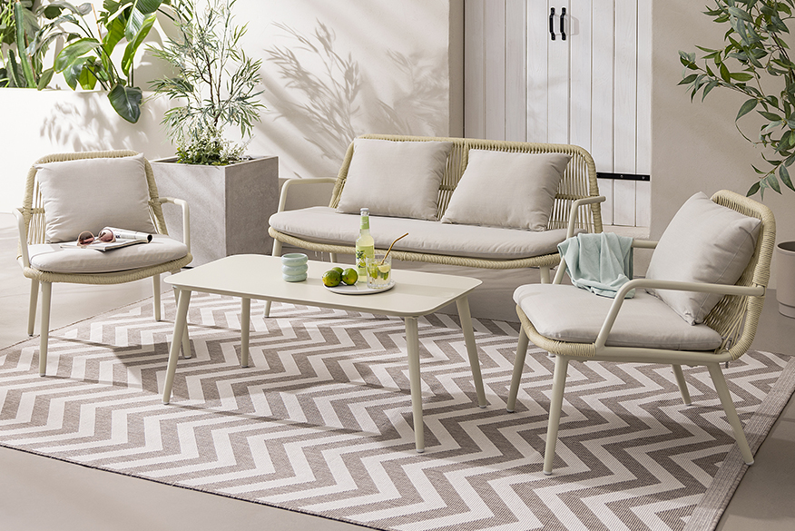 Beiges Outoorsofa im Rope Design mit passenden Sesseln und einem niedrigen Tisch, gemütlich hergerichtet mit cremefarbenen Polsterkissen und einem beige gezackten Teppich; daneben Pflanztöpfe und Gießkanne ebenfalls in Beige.