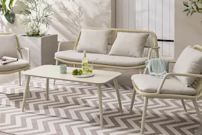 Canapé d’extérieur beige au design en corde, accompagné de fauteuils assortis et d’une table basse, avec des coussins d’assise couleur crème et un tapis beige à motif zigzag ; à côté, des pots de fleurs et un arrosoir également beige.