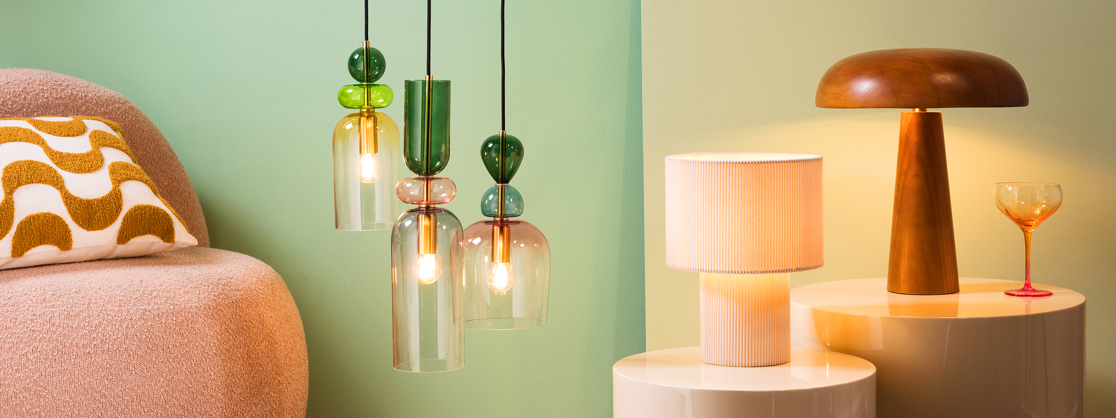 Mehrflammige Hängeleuchte aus Glas in bunten Farben, daneben eine Tischlampe aus Cordstoff sowie eine Mushroom Lamp aus Gummibaumholz.