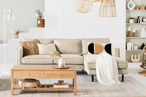 Salon avec des meubles des séries Fort Dodge et Taylor, de notre marque exclusive kollected : un canapé beige, une table basse avec motifs tressés, des suspensions en rotin, des coussins, tapis et plaids dans les tons beige et crème ainsi que des objets décoratifs sur une étagère murale.