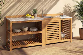 Cuisine d'extérieur en bois miel avec plan de travail aspect terrazzo clair, des étagères ouvertes et un évir intégré, sur une terrasse ensoleillée avec des plantes. Vaisselle bleue avec asperges et pain.