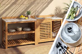 Outdoorküche aus warmem Holz mit heller Arbeitsplatte, offenen Regalen und integriertem Spülbecken auf einer sonnigen Terrasse mit Pflanzen. Blaues Geschirr mit Spargel und Brot.