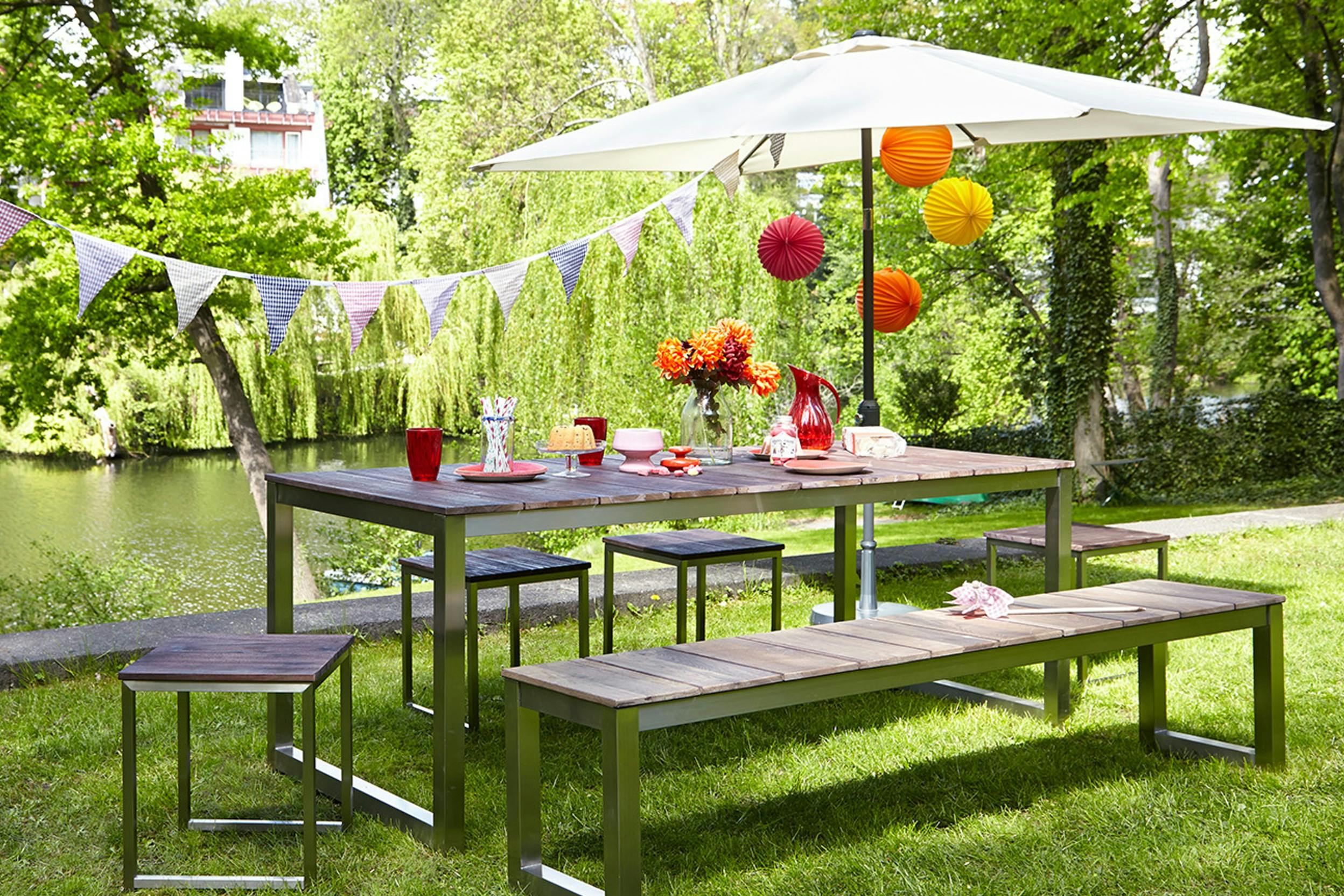 Sitzgruppe mit großem Tisch, Bank und Hockern auf einer grünen Wiese mit Sonnenschirm und Girlanden