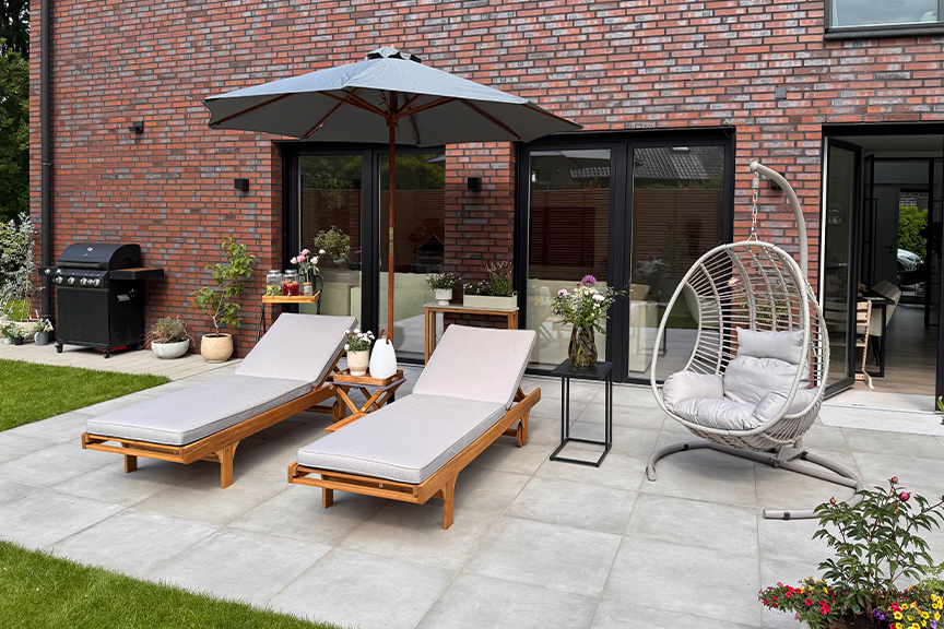 Schöne Terrasse mit hellbeigen Sonnenliegen, einem dunkelgrauen Sonnenschirm und einem Hängesessel.