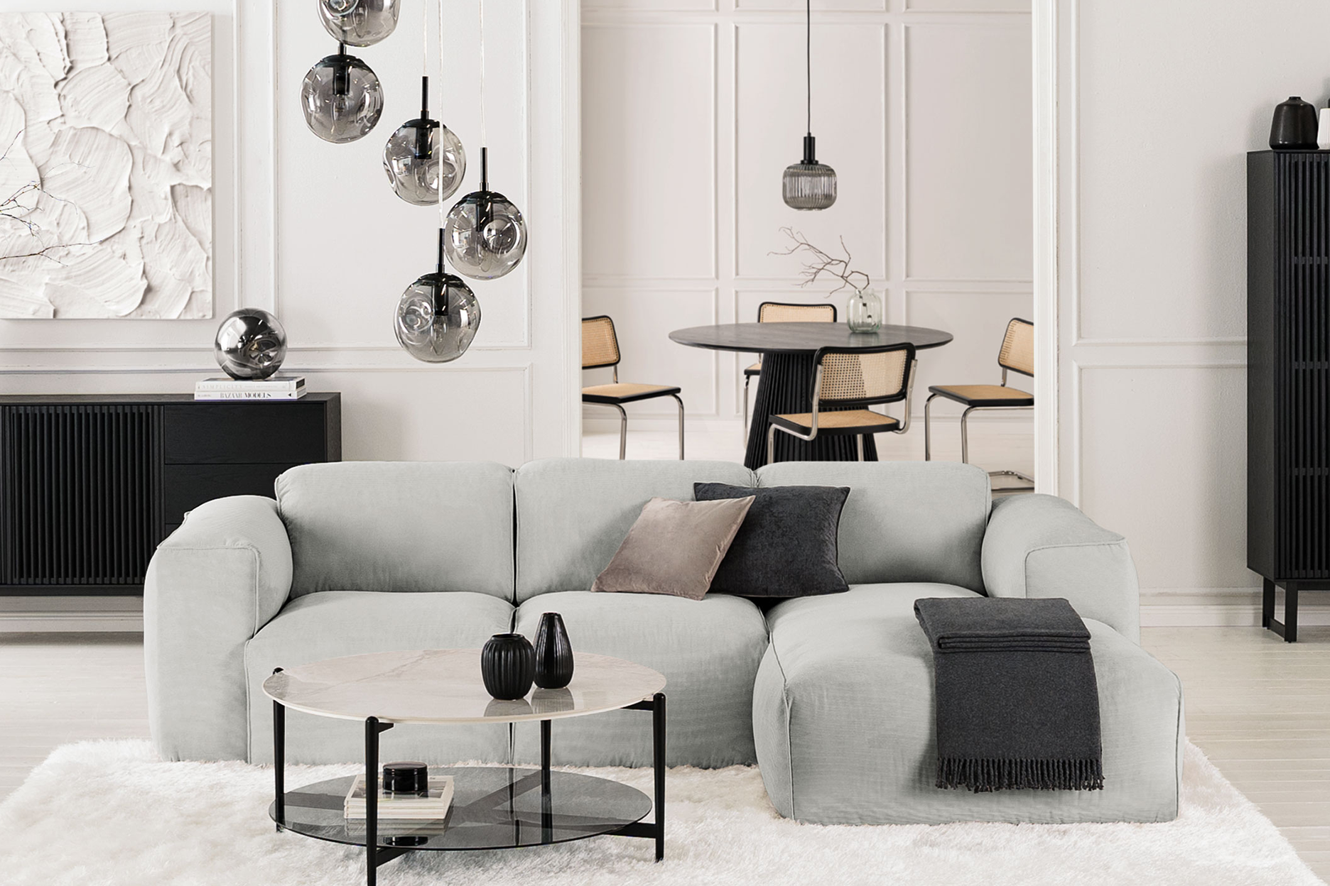 Modern eingerichtetes Wohnzimmer in unifarben Weiß sowie Grau und Schwarz, Sofa Hudson in der Mitte des Raumes, dazu skulpturale Leuchten aus Rauchglas, die schwarzen Stauraummöbel in Lamellen-Optik stammen aus der Serie Qara.