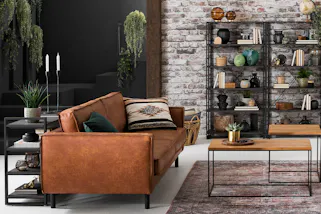 Wohnzimmer im Industrial-Stil mit braunen Ledersofas, zwei Couchtischen im coolen Holz-Metall-Mix und einem offenem Metallregal vor einer Backsteinwand.