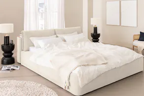Chambre minimaliste avec un lit blanc en velours côtelé crème, entouré de nombreux textiles aériens ; à côté, un tableau représentant un ciel parsemé de petits nuages blancs.