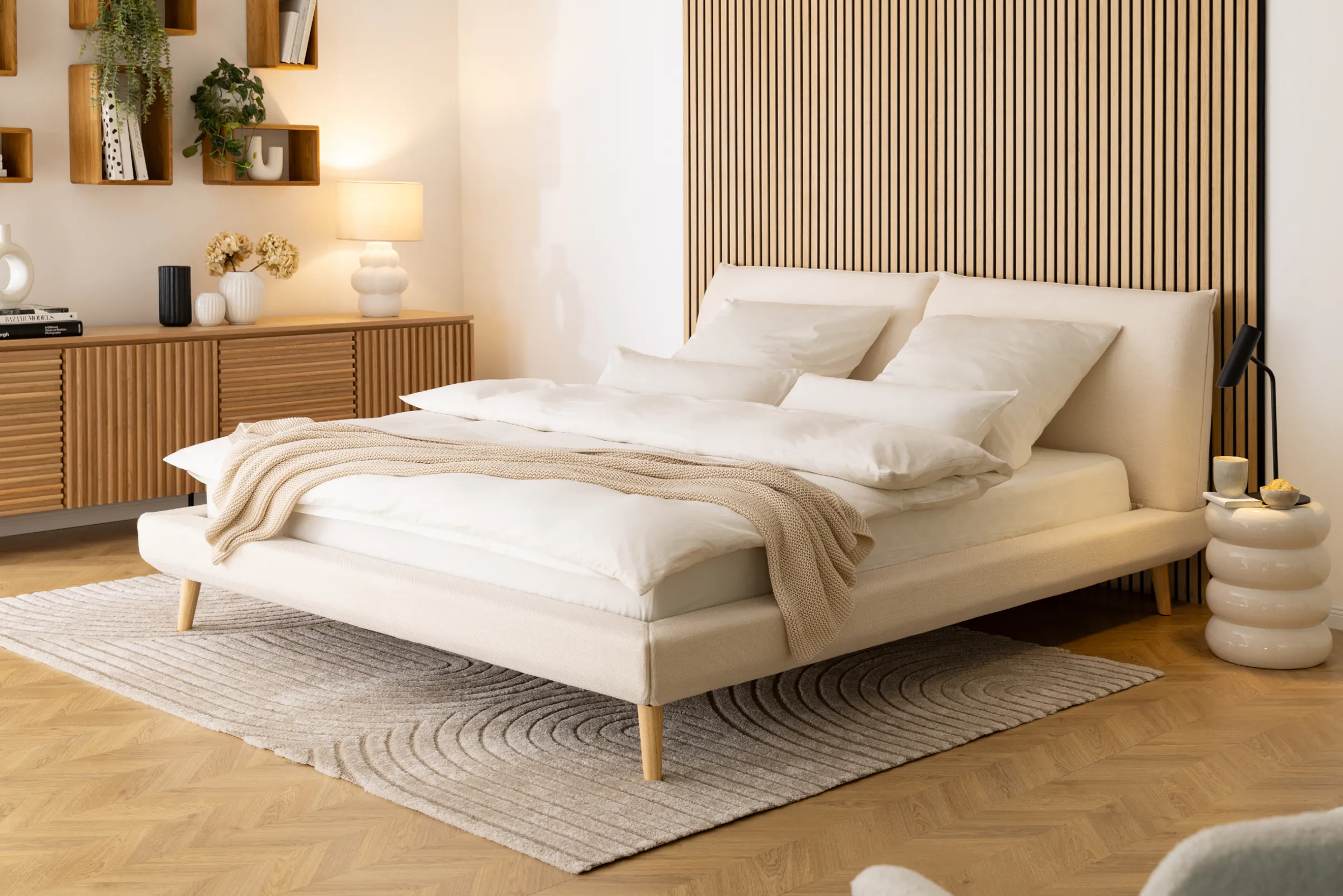 Innenaufnahme eines modernen, stilvollen Schlafzimmers. Ein beiges Doppelbett mit Holzbeinen steht auf Fischgrät-Holzboden. Ein großformatiger, rechteckiger Hochflorteppich (Modell Lioraen) mit einem subtilen, gewellten Kreismuster liegt zur Hälfte unter dem Bett und ragt großzügig am Fußende hervor. Im Hintergrund ist eine vertikale Holzlamellenwand, ein Sideboard und Wandregale mit Pflanzen zu sehen.