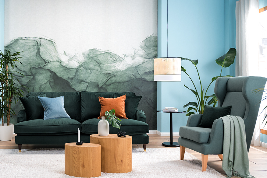 Wohnzimmer mit Samtsofa in Petrol, einem hellblauen Sessel und einem Retro-Sessel mit Wiener Geflecht, an der Wand eine blaugrüne Fototapete, dazu ein weißer Teppich, zwei Tische aus hellem Holz, ein schwarzer Beistelltisch, eine Pendelleuchte mit Wiener Geflecht sowie ein Sideboard, Deko und Wandbilder mit Tier- und Blumenmotiven.