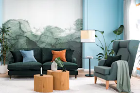 Wohnzimmer mit Samtsofa in Petrol, einem hellblauen Sessel und einem Retro-Sessel mit Wiener Geflecht, an der Wand eine blaugrüne Fototapete, dazu ein weißer Teppich, zwei Tische aus hellem Holz, ein schwarzer Beistelltisch, eine Pendelleuchte mit Wiener Geflecht sowie ein Sideboard, Deko und Wandbilder mit Tier- und Blumenmotiven.
