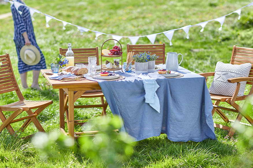 Gartenmöbelset aus Holz von BUTLERS, platziert auf einem grünen Rasen für ein Picknick: dazu Geschirr, Gläser und eine Etagere sowie ein weißes Kissen, eine blaue Tischdecke und eine weiße Wimpelkette.