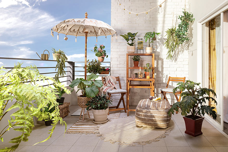 Balkon mit hellem Boden und heller Wand, der mit klappbaren Balkonmöbeln aus Holz sowie beigefarbenen Outdoor-Teppichen, Textilien und Sonnenschirm im Boho-Stil gestaltet ist und mit vielen Pflanzen begrünt wird