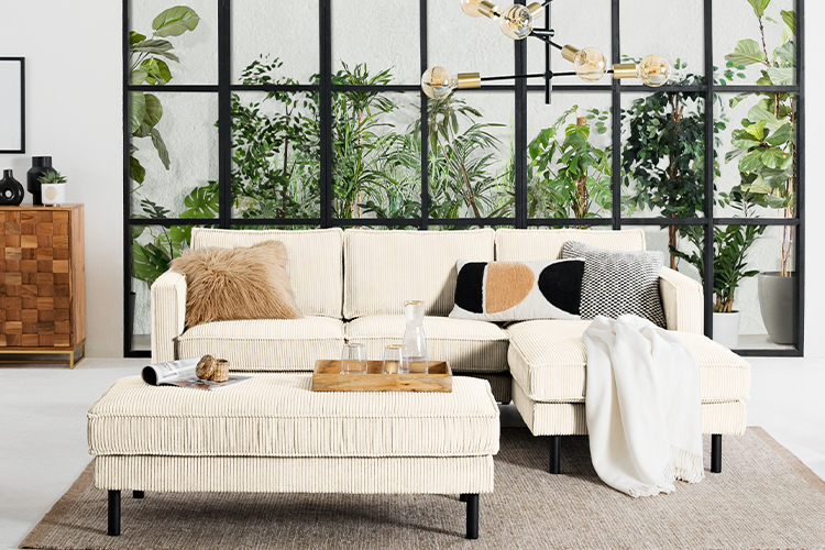 Ecksofa Fort Dodge und XL Polsterhocker Fort Dodge in einem abwechslungsreich eingerichteten Wohnzimmer im Boho-Stil, dazu gemütliche Textilien, ein Rattansessel, eine Industrial-Lampe sowie ein Sideboard aus Massivholz, im Hintergrund ein Fenster zu einem Lichthof mit vielen tropischen Zimmerpflanzen.