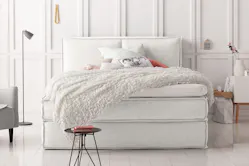 Weißes Boxspringbett mit kuscheliger Tagesdecke vor weißer Wand