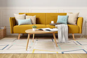 Wohnzimmer mit gelbem Retro-Sofa mit beigen und blauen Kissen, buntem Teppich und einem Couchtisch