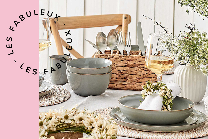 Table en bois dressée avec chemin de table blanc, vaisselle en céramique bleu-gris, range-couverts et assiette de service en rotin ainsi qu'une décoration avec des fleurs séchées dans un vase et une couronne de fleurs ; à côté, un ensemble de sièges en bois dans un jardin ensoleillé avec une table de petit-déjeuner et un arrosoir en fonte au premier plan.