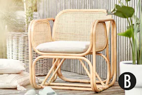 Outdoor-Sessel bespannt mit natürlichem Rattangeflecht auf einer sonnigen Terrasse, dazu ein Jute-Teppich und Baumwollkissen mit Quasten und anderen Applikationen.