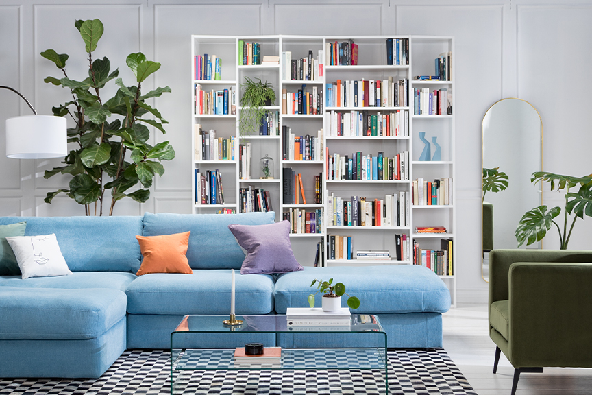 Wohnzimmer mit hellblauem Sofa, bunten Kissen, einem Glastisch sowie einem grünem Samtsessel, dazu eine Bogenlampe mit weißem Schirm, ein weißes Regal und ein schwarz-weiß karierter Teppich.