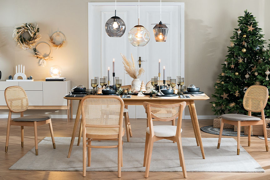 Esszimmer mit Möbeln von Studio Copenhagen: ein heller Esstisch aus Holz mit Rattanstühlen, ein weißes Sideboard, ein grauer Sessel sowie Pendelleuchten aus Glas; dazu geschmückte Weihnachtsbäume, weihnachtliche Tischdeko, schwarze Geschirrsets und Trinkgläser, schwarze und weiße Vasen mit Pampasgras sowie weitere Deko in Schwarz und Weiß.