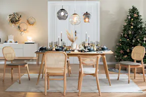 Esszimmer mit Möbeln von Studio Copenhagen: ein heller Esstisch aus Holz mit Rattanstühlen, ein weißes Sideboard, ein grauer Sessel sowie Pendelleuchten aus Glas; dazu geschmückte Weihnachtsbäume, weihnachtliche Tischdeko, schwarze Geschirrsets und Trinkgläser, schwarze und weiße Vasen mit Pampasgras sowie weitere Deko in Schwarz und Weiß.