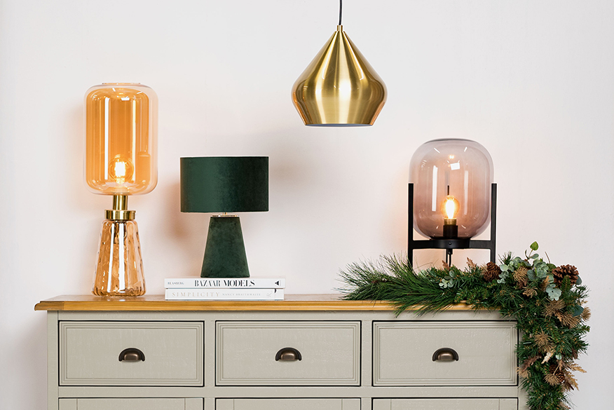 Kommode mit einer goldgelben Tischleuchte aus Glas, einer Tischleuchte aus grünem Samt, einer Pendellampe aus Messing sowie einer Tischleuchte mit schwarzem Rahmen und rauchfarbener Glaskugel. Daneben eine Stehlampe im Industrial-Stil aus schwarzem Metallgeflecht.