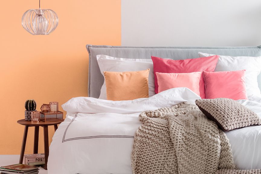 Hellgraues Boxspringbett Avellino by Mørteens kombiniert mit Kissen und Wandfarbe in der Pantone-Farbe 2024 „Peach Fuzz“ sowie weiteren Textilien, Leiterregal mit Pflanzen, Beistelltisch und kupferfarbener Deckenlampe.