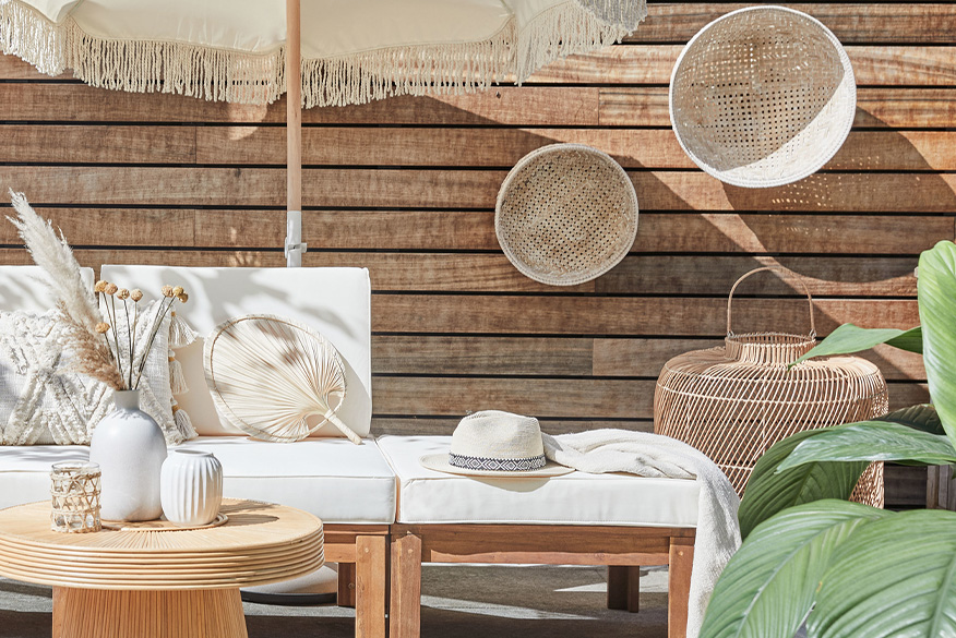 Outdoor-Loungemöbel der Serie LEXI by BUTLERS aus hellem Akazienholz mit weissen Polstern, dazu ein passender Tisch dekoriert mit weissen Vasen, ein weisser Sonnenschirm mit Fransen, ein Outdoor-Teppich und Pouf aus Jute sowie Wanddeko, Pflanzenkorb und Windlicht in Flechtoptik.