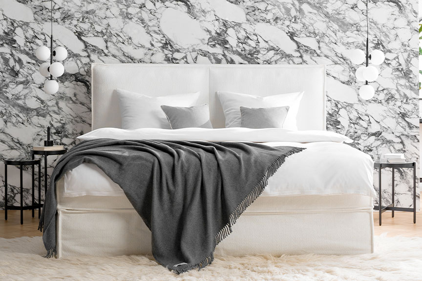 Weißes Boxspringbett KINX der home24 Exklusivmarke Studio Copenhagen vor einer Wand mit moderner Marmortapete, daneben schlanke Beistelltische, Hängelampen und ein Stuhl für abgelegte Kleidung mit Wiener Geflecht.