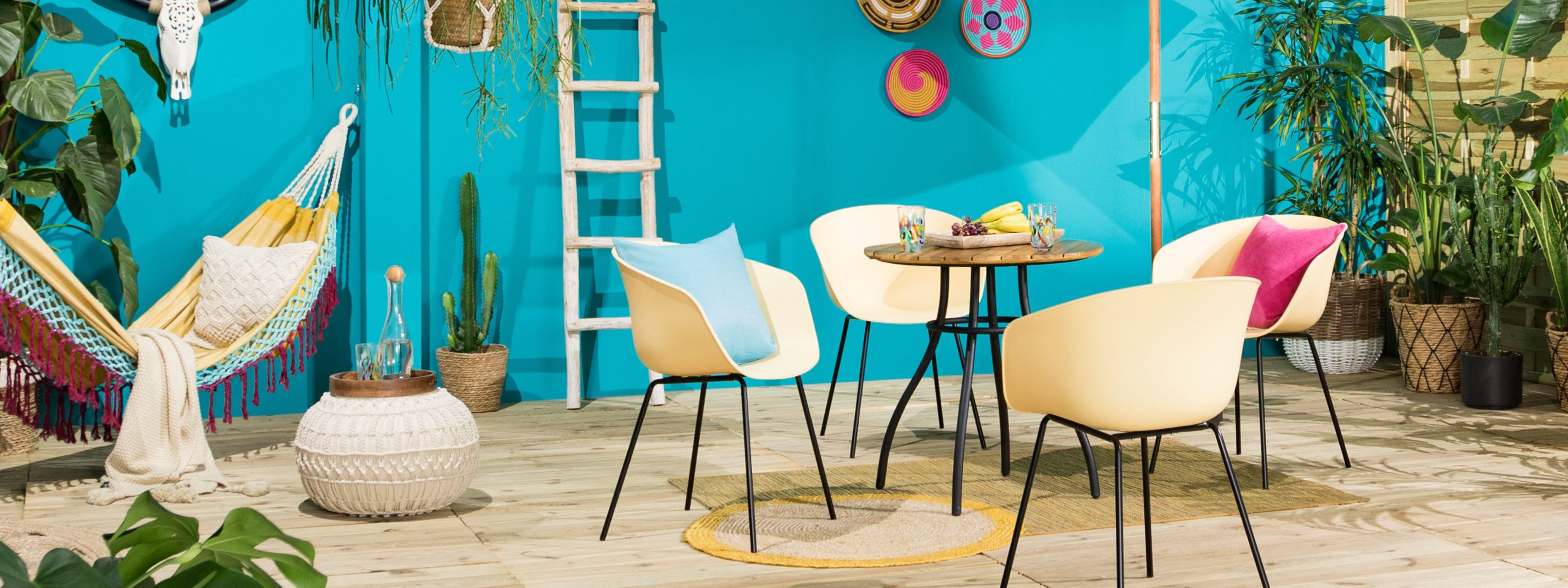 Terrasse mit türkisblauer Wand, davor ein Gartentisch im Industrial-Stil mit hellgelben Schalenstühlen, dazu ein beiger Strickpouf, ein weißer Sonnenschirm, eine bunte Hängematte und Wandteller sowie viele Pflanzen in Körben und Blumenampeln.