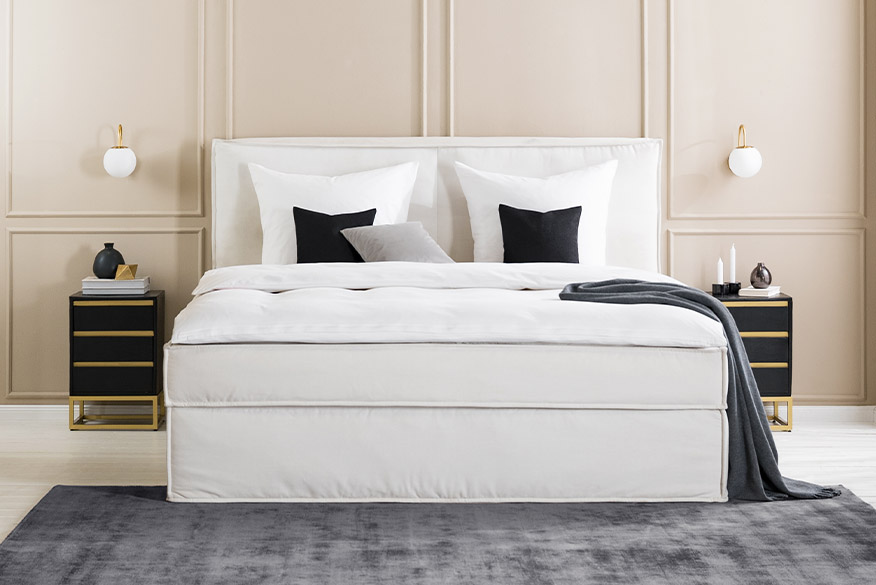 Einrichtungsbeispiel für Hotels: Schlafzimmer mit einem beigen Boxspringbett und Nachttischen in einer eleganten Schwarz-Gold-Kombination, dazu schwarze und weiße Deko und viele Textilien.