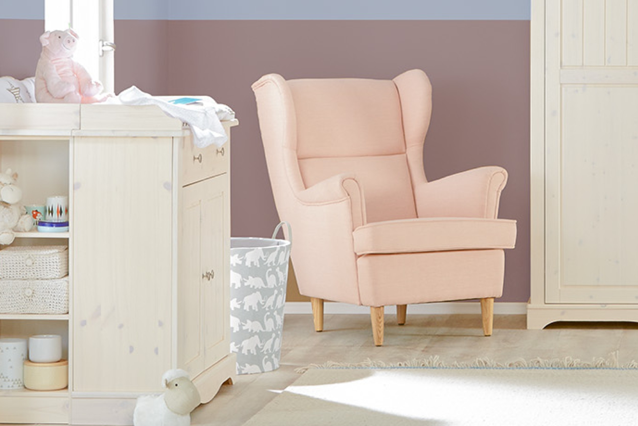 Babyzimmer mit hellen Möbeln sowie rosafarbener Ohrensessel stehend vor gestrichener Wand