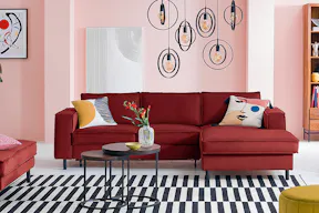 Rotes Sofa inkl. Polsterhocker der home24 Exklusivserie Fort Dodge by kollected mit bunten Kissen auf einem schwarz-weiß gestreiften Teppich. Dazu zwei runde Beistelltische aus dunklem Holz und Metall im Industrial-Stil, ein gelber Pouf, ein schwarzer Stuhl mit Wiener Geflecht und gelbem Kissen, mehrere schwarze Pendelleuchten, ein Kandinsky-Wandbild und Highboard Deva aus Holz und Metall.