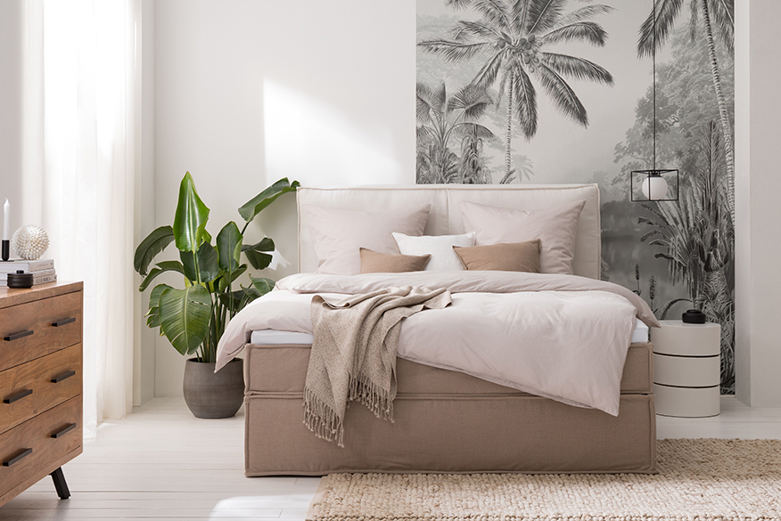 Schlafzimmer in Cremefarben mit einem Boxspringbett in Beige und heller Bettwäsche, dazu eine Kommode und Pendelleuchte im Industrial-Stil.