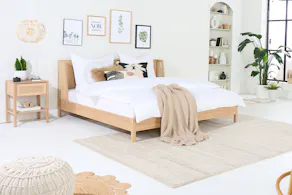 Chambre à coucher avec des meubles en chêne clair de la série Taylor, by kollected, une marque exclusive home24 : un lit, une table de nuit, une chaise à bascule, un bureau avec chaise de bureau, ainsi que de nombreuses plantes vertes, des tapis, des textiles et un pouf en tricot. Tous les produits Taylor ont en commun l'alliance du chêne clair et du rotin.