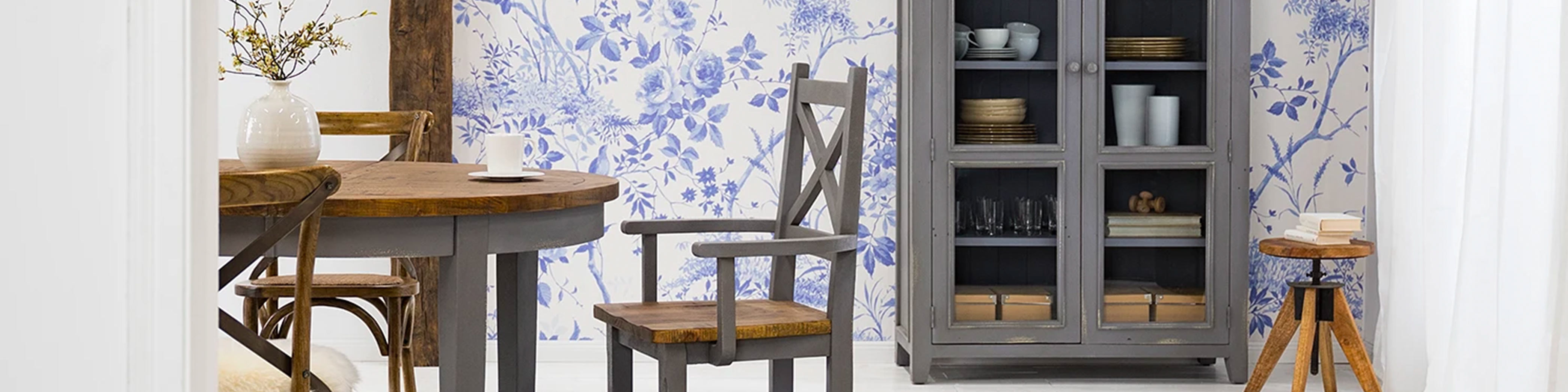 Meubles de salle à manger gris de style maison de campagne devant du papier peint fleuri bleu et banc.