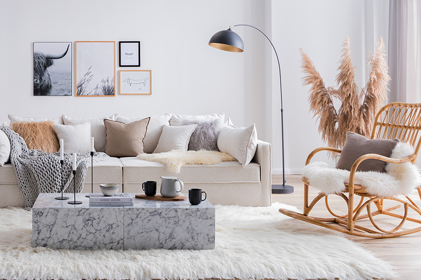 Woonkamer in de hygge-stijl met een beige bank van bouclé, een rotan schommelstoel en een stijlvolle marmeren salontafel, plus veel kussens, een plaid, een pluizig wit vloerkleed, een zwarte booglamp, een dressoir van licht hout, pampagras, accessoires in zwart en rotan en foto's aan de muur.
