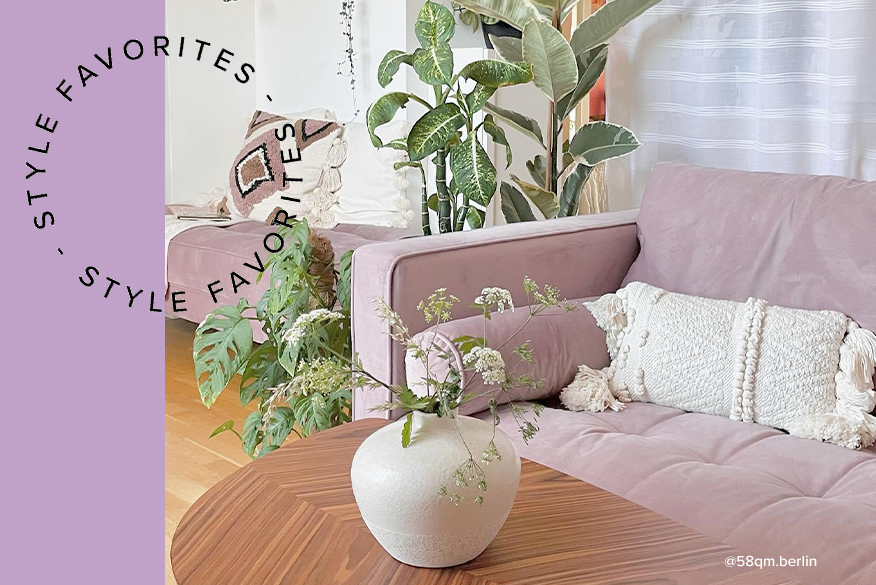 Lilafarbenes Sofa mit weißen Kissen im Boho-Style, dazu viele Grünpflanzen; daneben eine Glasvase mit rosa Pfingstrosen, dahinter eine Blumentapete.