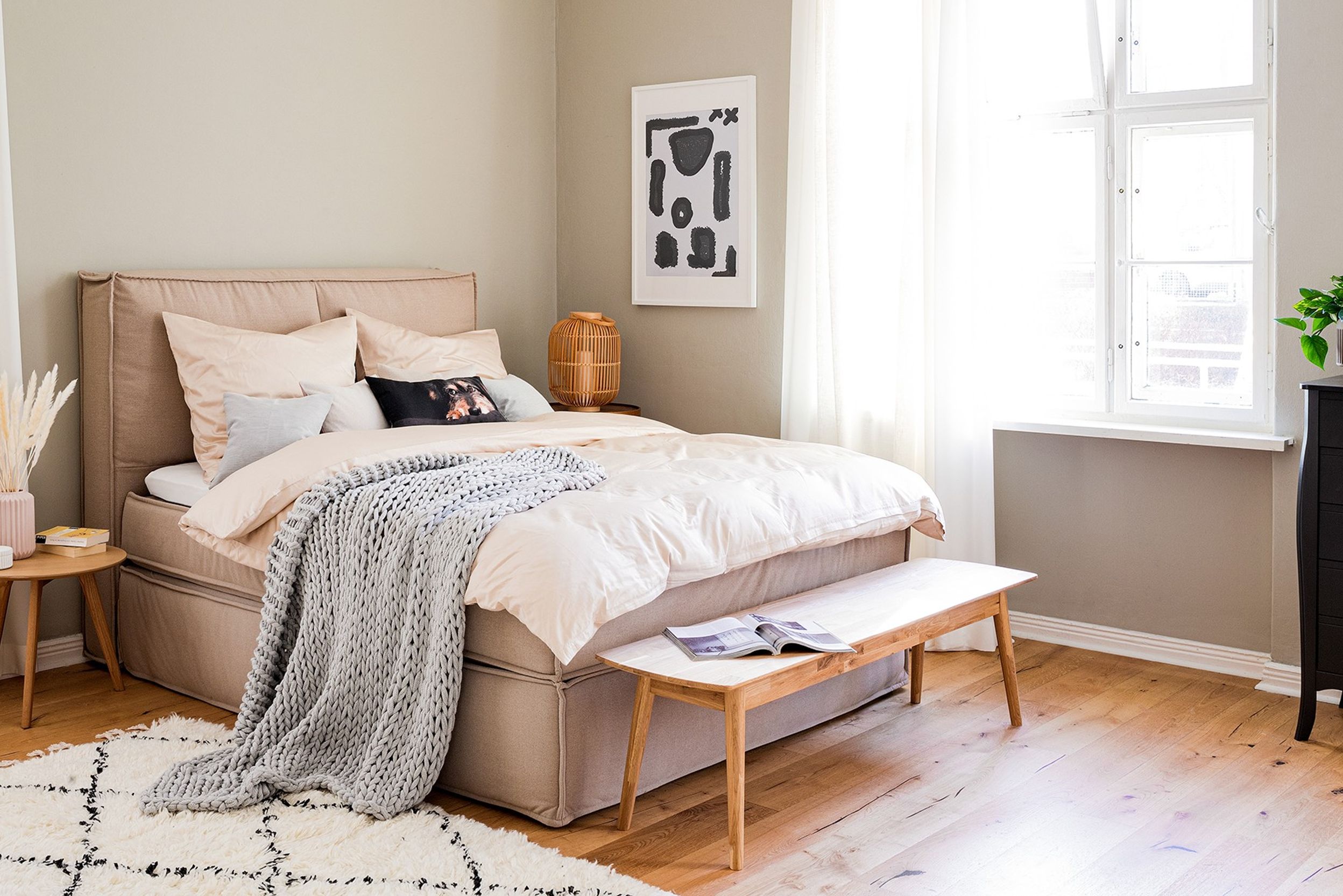 Chambre aux tons clairs, des variations de beige et blanc, avec déco, fenêtre aux rideaux blancs, cadre photo noir et blanc, lit beige, linge de lit beige et plaid gris clair