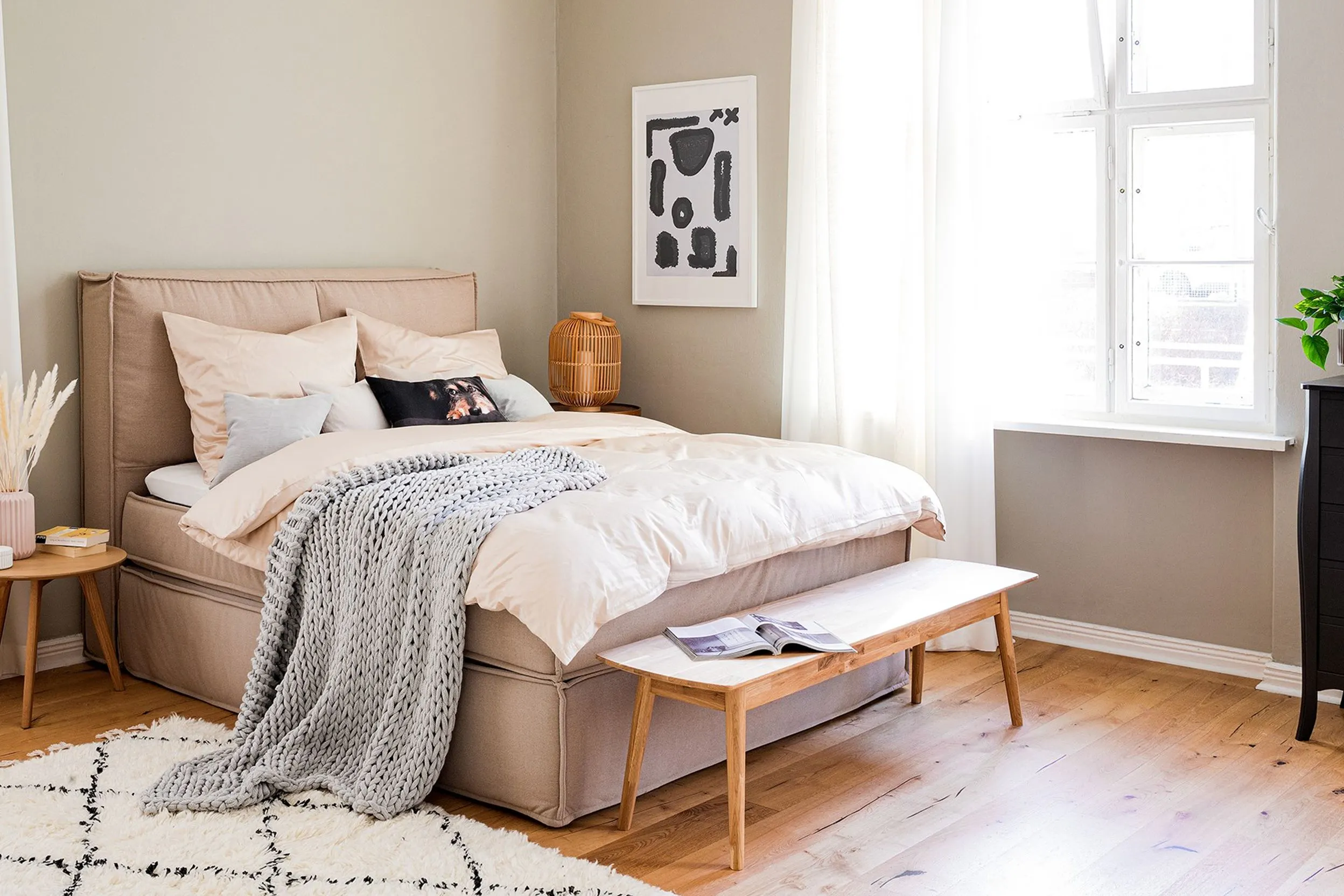 Chambre aux tons clairs, des variations de beige et blanc, avec déco, fenêtre aux rideaux blancs, cadre photo noir et blanc, lit beige, linge de lit beige et plaid gris clair