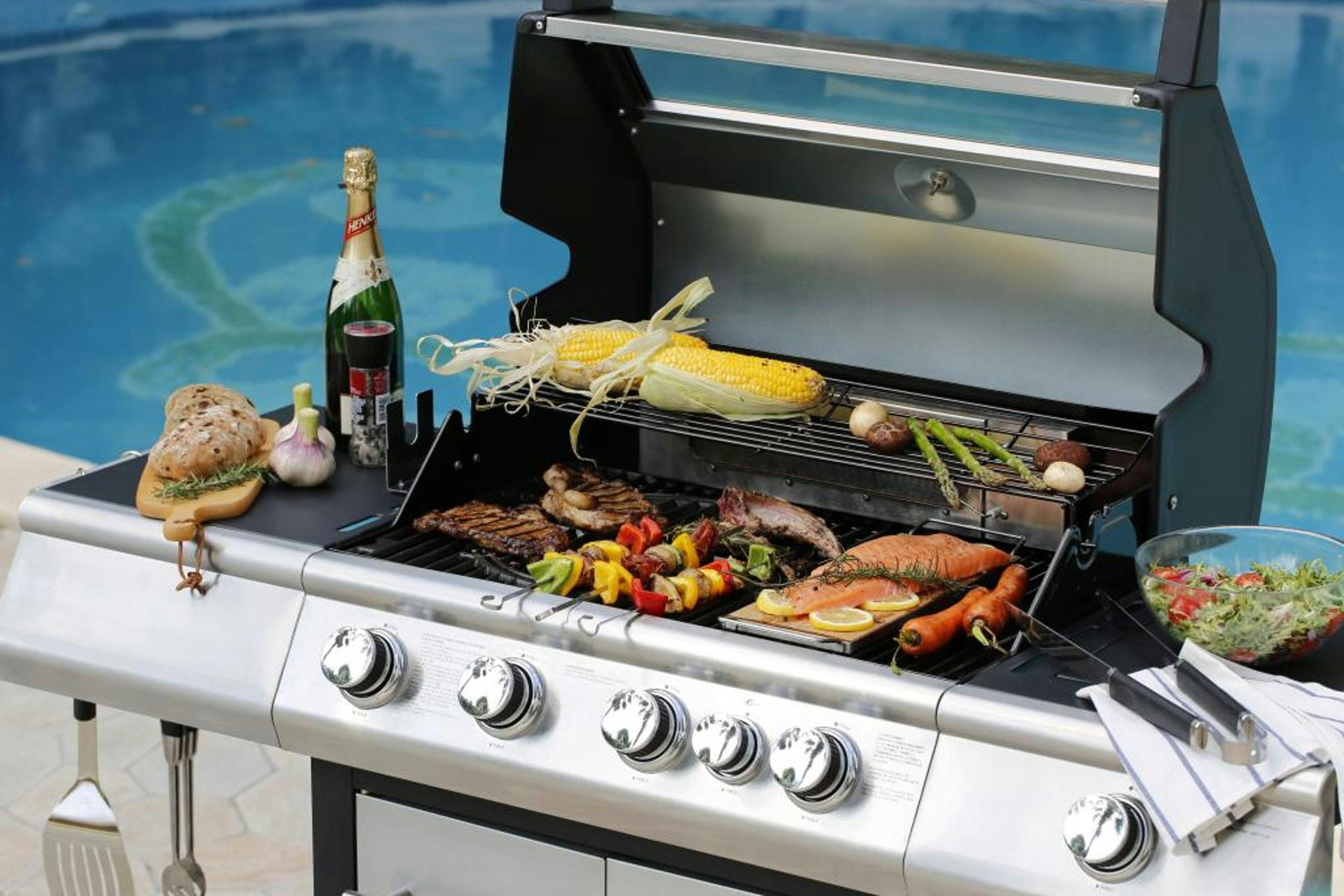 Großer Gasgrill mit Gemüse, Fisch und Fleisch vor einem blauen Pool