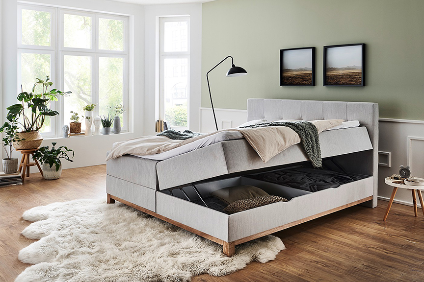 Hellgraues Polsterbett mit zwei Bettkästen für mehr Stauraum im Schlafzimmer, welches vor mintgrüner Wand auf einem Holzfußboden steht