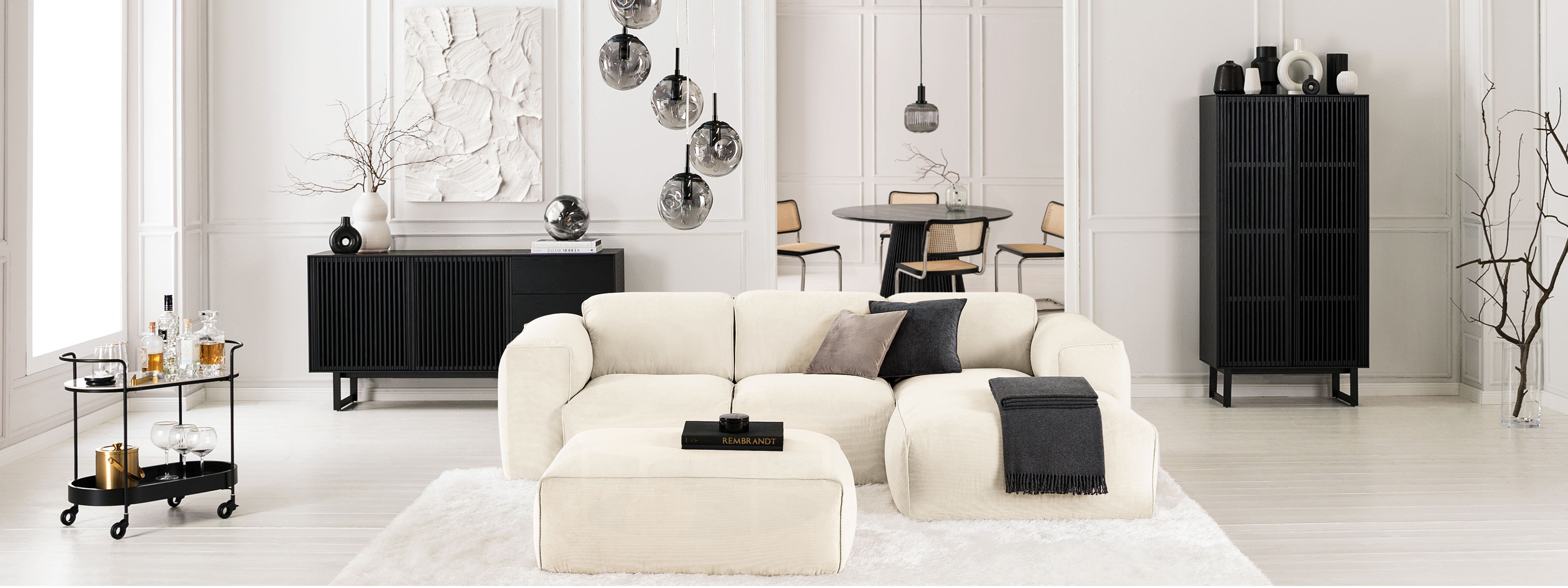 Salon noir et blanc avec canapé blanc Hudson de chez Studio Copenhagen sur un tapis fluffy blanc, avec meubles noirs posés devant des murs blancs et présentant des objets déco noirs et blancs.