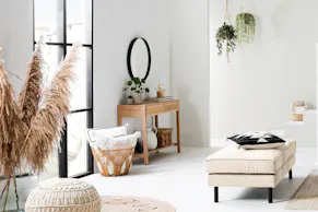 Tabouret capitonné beige avec des pieds noirs, un bureau en cannage, un miroir noir au-dessus, un panier avec des coussins à côté. En outre, de l'herbe de la pampa et un pouf en tricot pour la décoration.