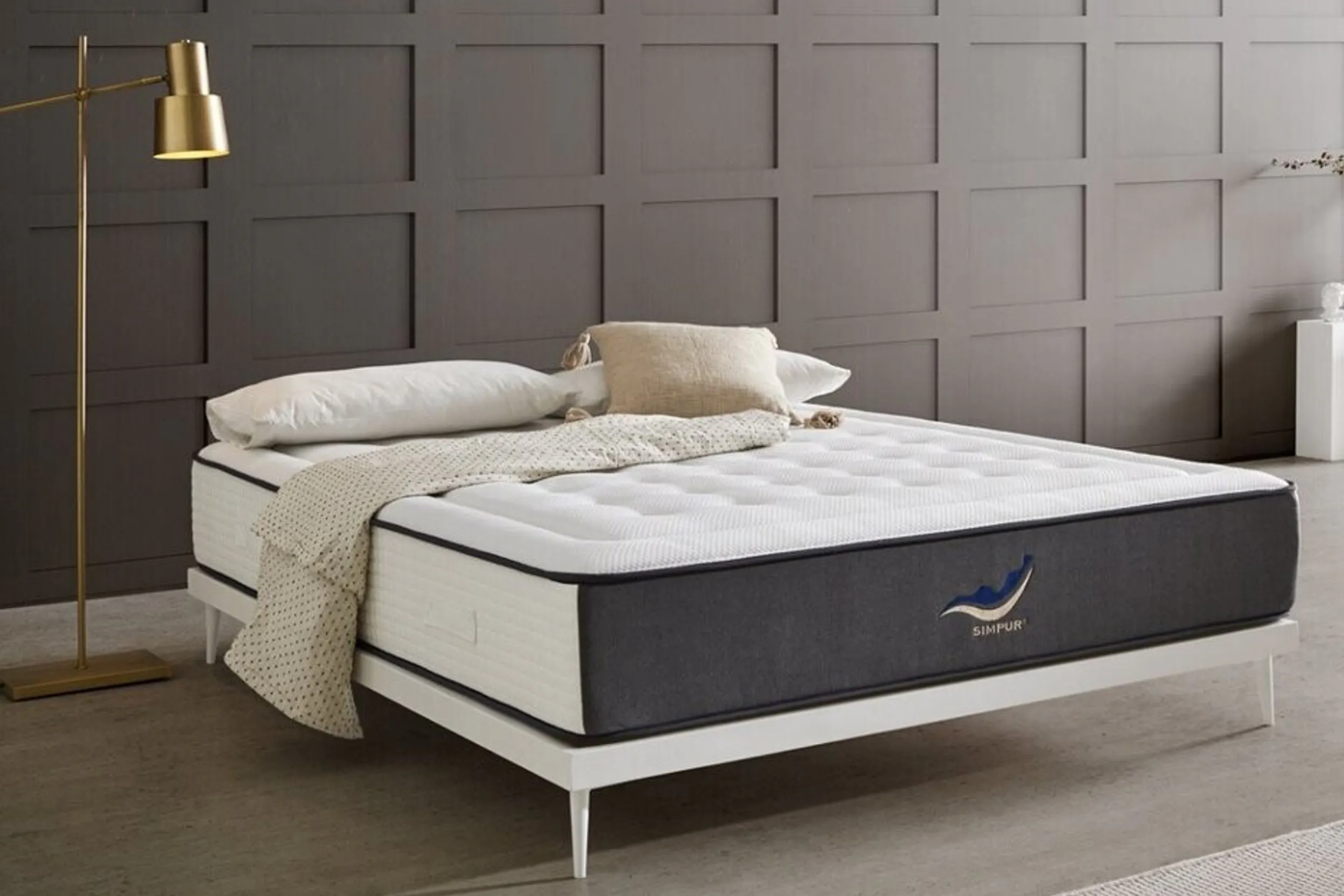 Weißes Bettgestell mit dicker Matratze und goldener Stehlampe vor einer grau getäfelten Wand