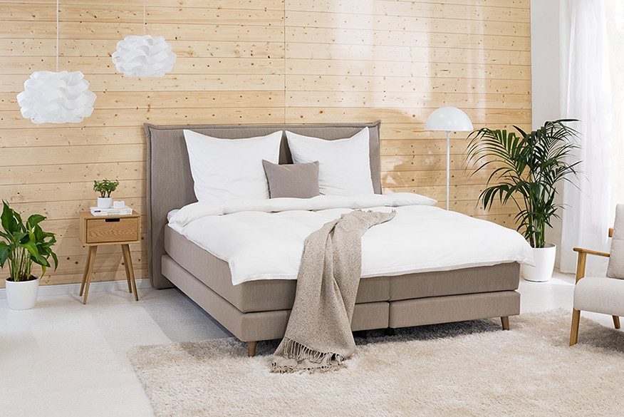 Schlafzimmer in Neutralfarben mit einem Bett, Kissen, Decke und Teppich in Taupetönen, dazu weiße Bettwäsche, ein weißes Sideboard, ein Nachttisch aus Holz, ein Sessel im Retro-Stil, Grünpflanzen, eine weiße Stehleuchte sowie weiße Pendelleuchten.