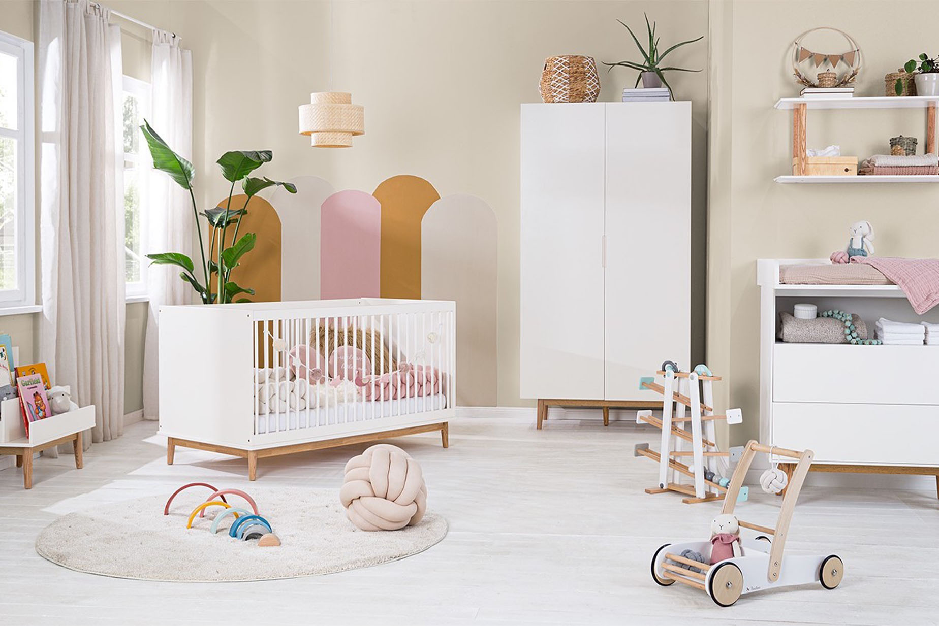 Chambre bébé avec lit à barreaux blanc et armoire de style scandinave, tapis, coussins et linge de lit dans les tons crème, tour de lit tressé rose et arche d'activité en bois dans les tons pastel.