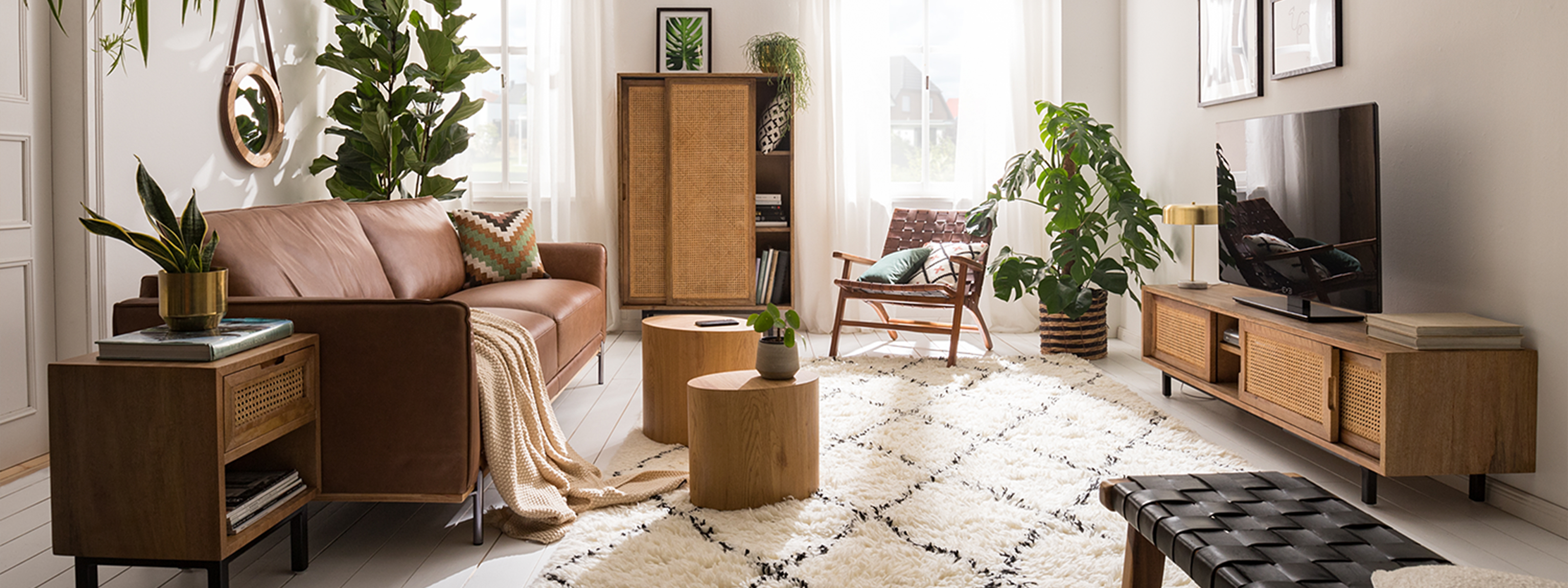 Wohnzimmer im Boho-Style mit einem Ledersofa, Holzmöbeln mit Wiener Geflecht, Rattan, vielen Pflanzen, Textilien und Deko aus Messing