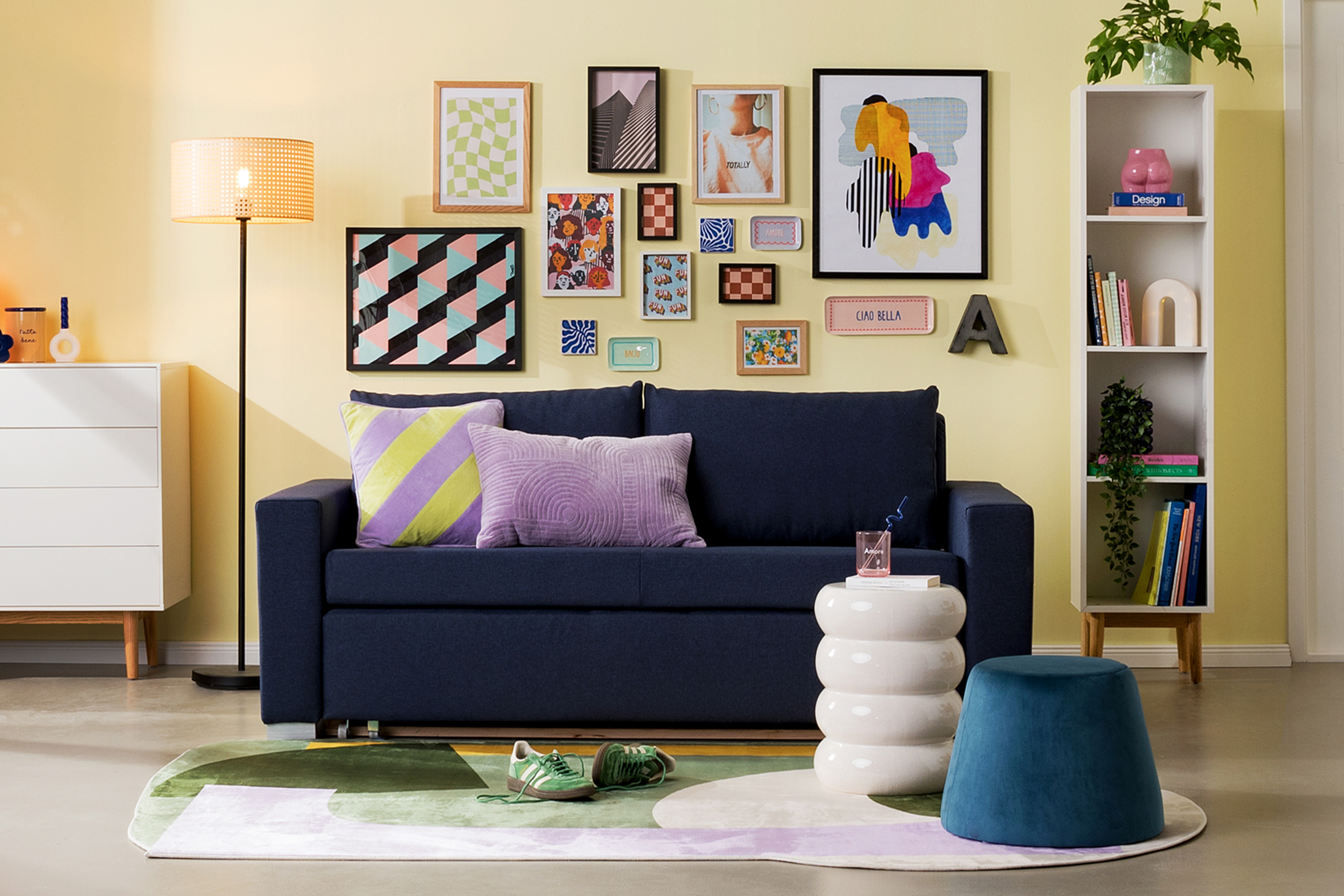 Wohnzimmer oder WG-Zimmer mit blauem Schlafsofa, einer weißen Kommode im Skandi-Stil, einem weißen Regal, einem blauen Pouf, einer Rattan-Stehlampe, einem weißen Wandschreibtisch, einem Armlehnenstuhl in Senfgelb, einem Spiegel und viel bunter Deko.