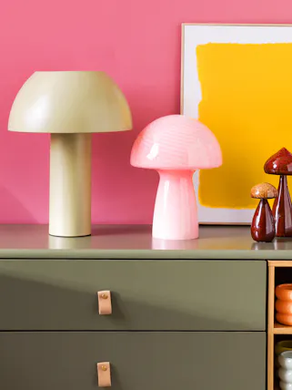 Mehrere bunte Tischlampen in Pilzform auf einem dunkelgrünen Sideboard vor pinkfarbener Wand.