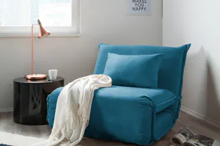 Blauer Schlafsessel mit weißer Decke, dazu eine kupferfarbene Tischleuchte auf einem schwarzen Beistelltisch.